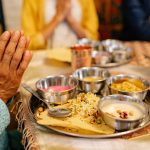 A close-up image of a traditional Rice meal served on a metal thali, featuring rice, papad, and small bowls with various curries and desserts, with hands clasped in prayer in the foreground.