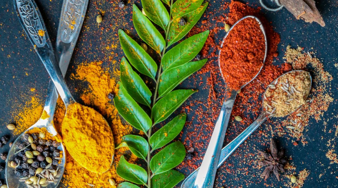 A colorful display of spices and herbs on a dark surface, including curry leaves, turmeric, chili powder, and star anise in ornate metal spoons.